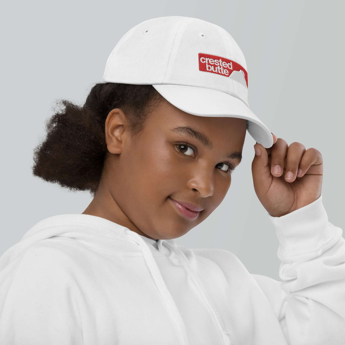 Crested Butte Youth Baseball Cap worn by a girl in a white hoodie, promoting official ski resort apparel by Mtns Co.